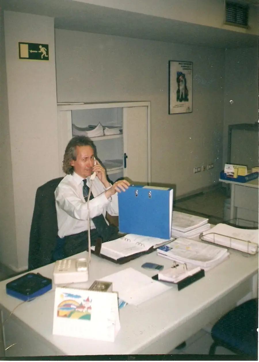 A man in an office sits at a desk talking on the phone, holding a blue binder. The desk is cluttered with papers, folders, a telephone, and a calendar. Shelves and office equipment are visible in the background.