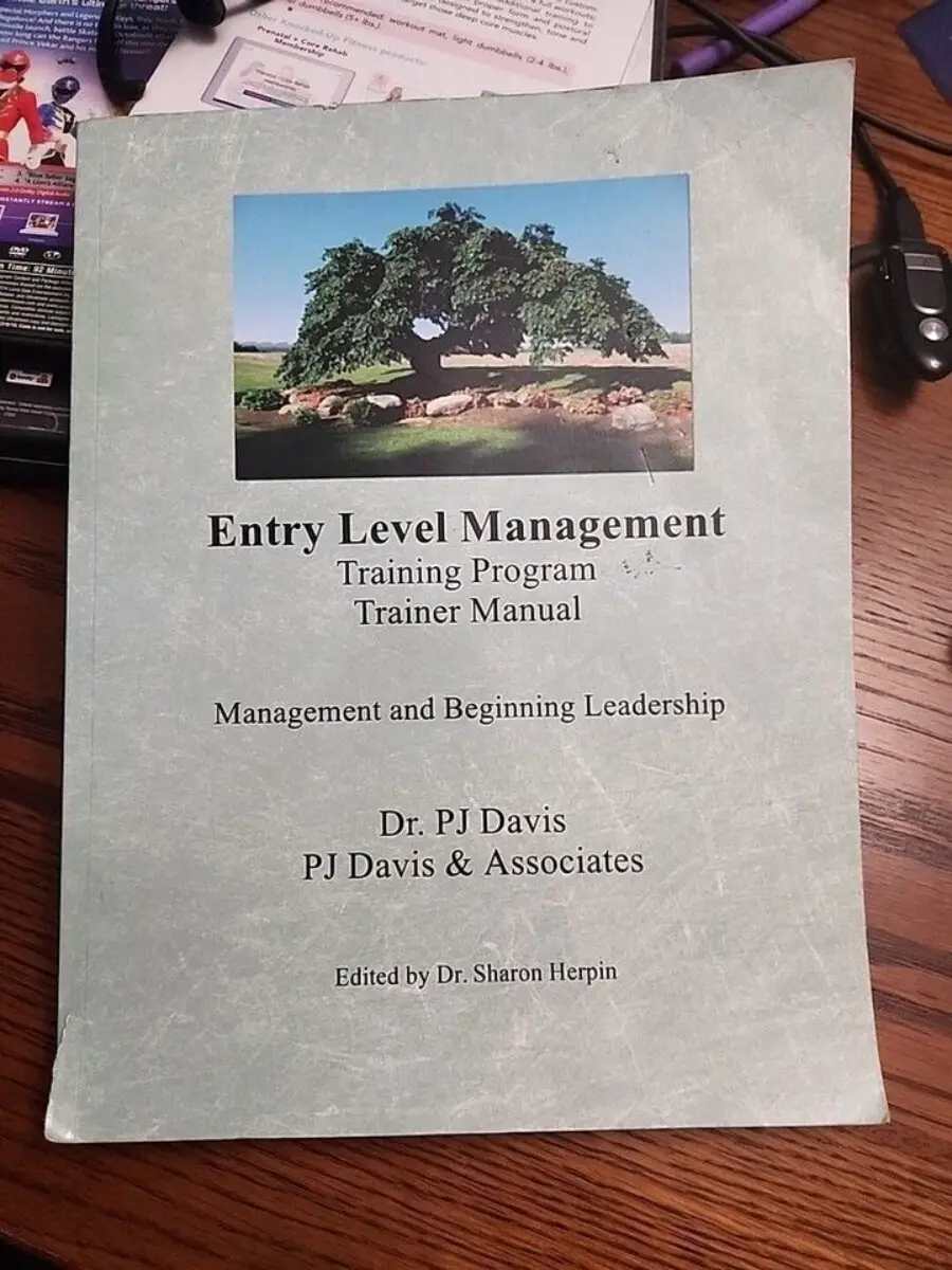 A book titled "Entry Level Management: Training Program Trainer Manual" by Dr. PJ Davis, PJ Davis & Associates, edited by Dr. Sharon Herpin, is on a desk. The cover features a large tree and some office items are visible nearby.