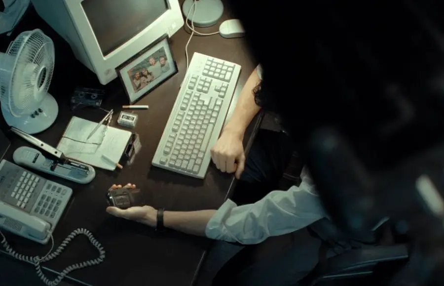 A person sits at a cluttered desk with a vintage computer, keyboard, phone, small fan, notepad, cassette tapes, and a framed photo. The person holds a cassette recorder in their hand. The scene has a retro, nostalgic feel.