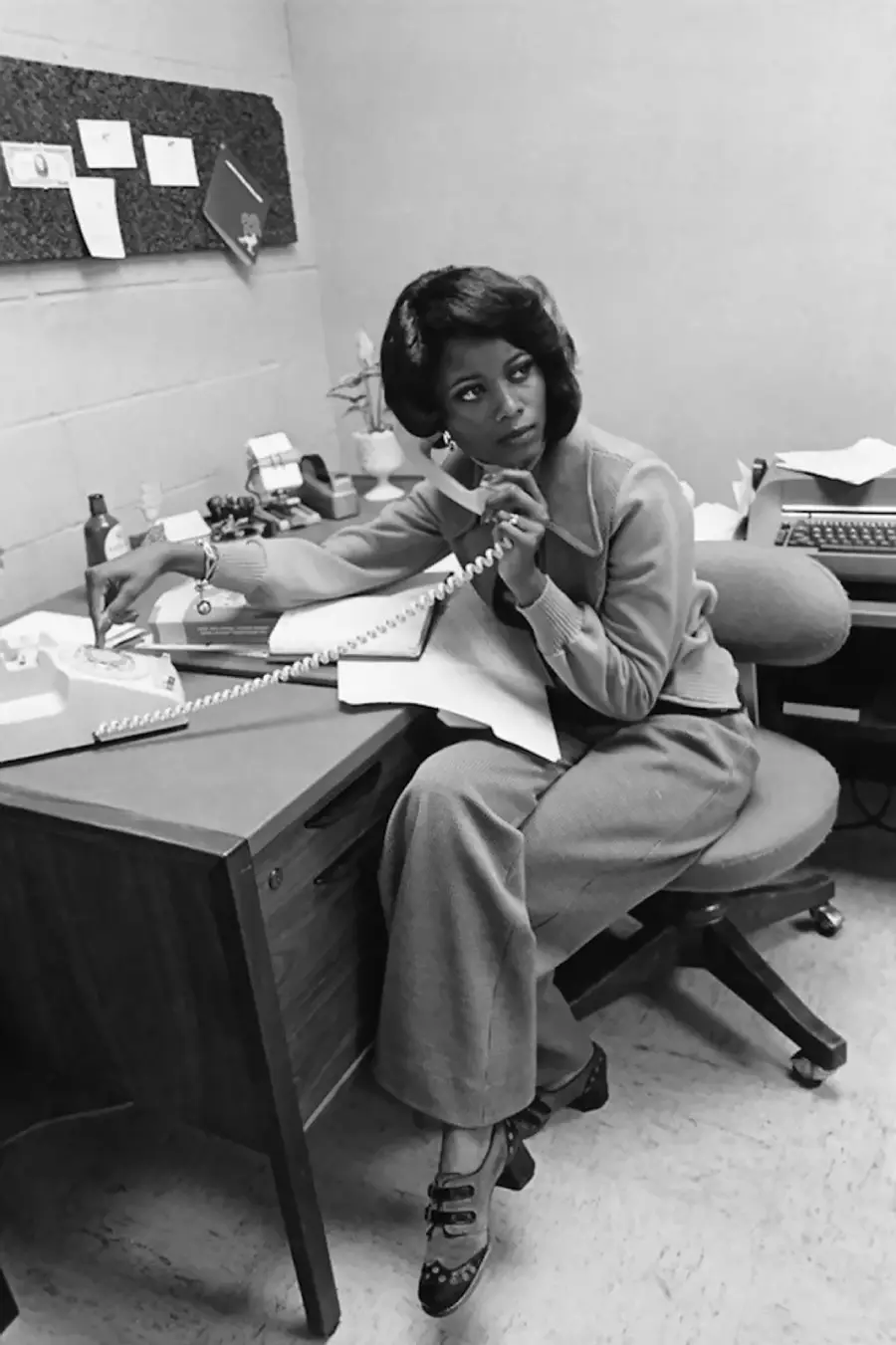 A woman sits at an office desk talking on a corded telephone, holding a pen and writing on papers. She wears a suit and heels, with office supplies and papers on the desk in a 1970s-style setting.