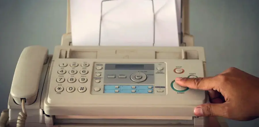 A person’s hand presses the start button on a beige fax machine, which has paper loaded in the tray and a telephone handset attached on the left side.