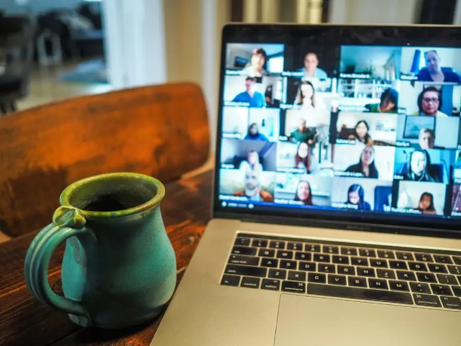A green ceramic mug sits on a wooden table next to a laptop. The laptop screen displays a video call with many participants in a grid layout. The background is slightly blurred.