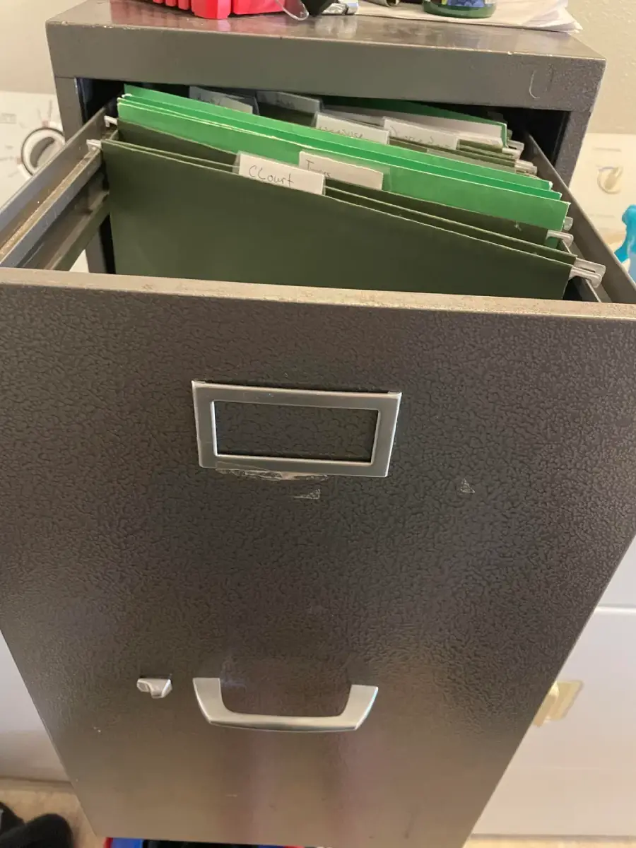 A partially open black metal file cabinet drawer containing several green hanging file folders with labeled tabs, set in an indoor setting with some objects visible in the background.