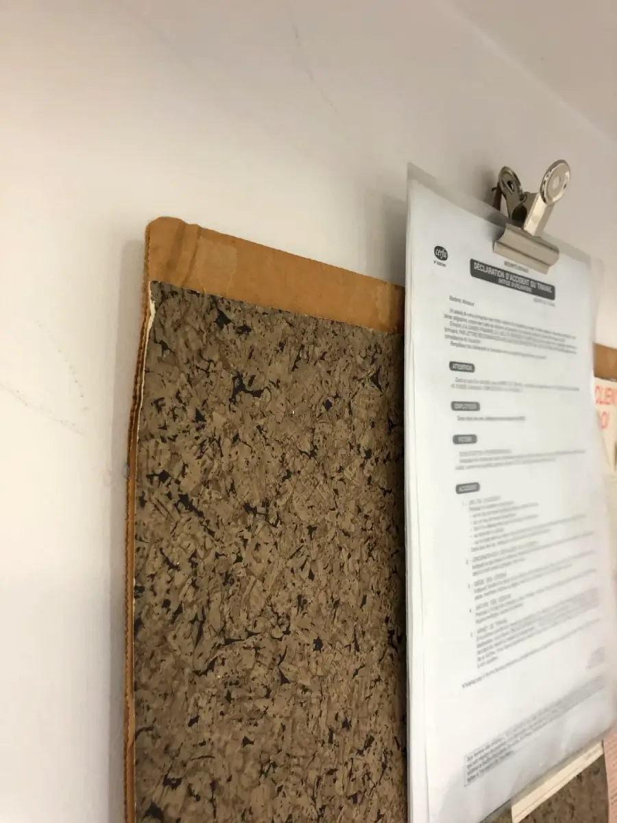 A close-up of a corkboard with a brown cardboard edge and a metal clip holding a printed document against a white wall. The document appears to be an official form or notice.