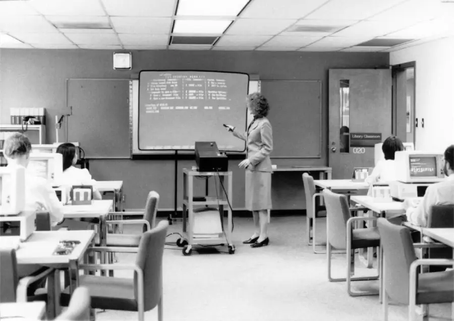 A woman stands beside an overhead projector, pointing at a screen in a classroom with students seated at desks, facing her. The scene appears to be from the 1980s, with vintage office furniture and computer equipment.