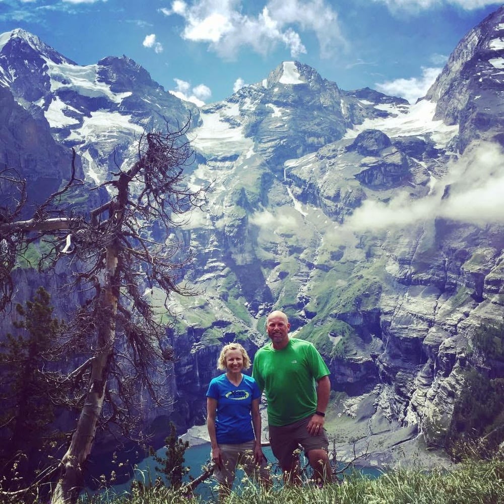 Two people stand smiling in front of a scenic mountain landscape with snow-capped peaks, green slopes, a lake below, and a partly cloudy blue sky. A tree leans to the left in the foreground.