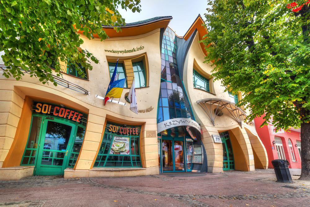 A whimsical, warped building with wavy walls and windows, featuring a glass entrance and “SO! COFFEE” signs. Trees line the sidewalk in front, and colorful walls add to the playful architectural style.
