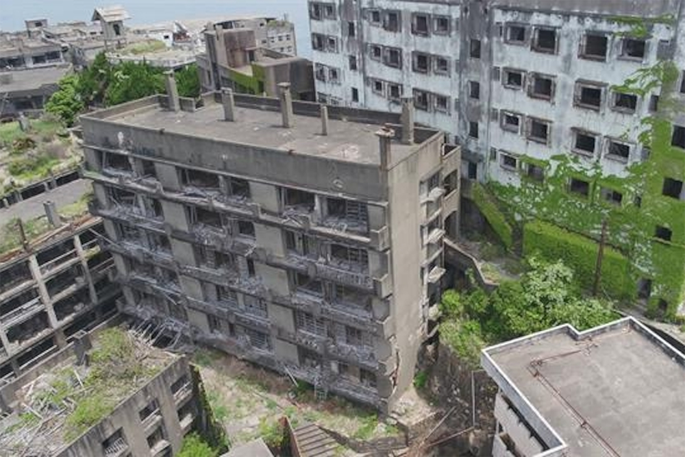 Aerial view of abandoned, decaying concrete buildings with broken windows and overgrown vegetation. The scene looks deserted and rundown, suggesting long-term neglect.