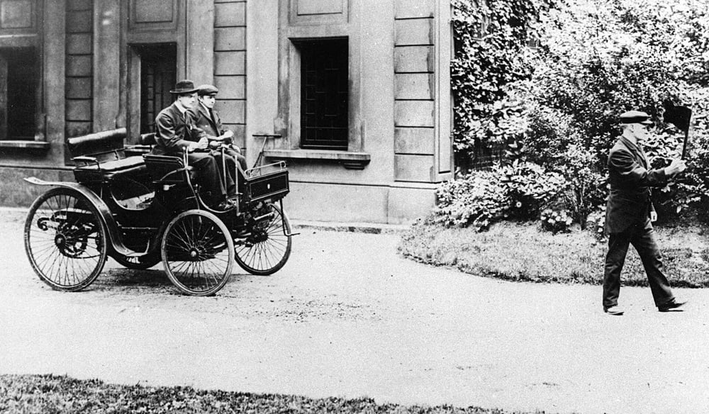 Two people sit in an early motor car with large wheels and no roof, while a man in uniform walks ahead of the vehicle holding a flag. The scene is outside a building with tall windows and surrounding greenery.