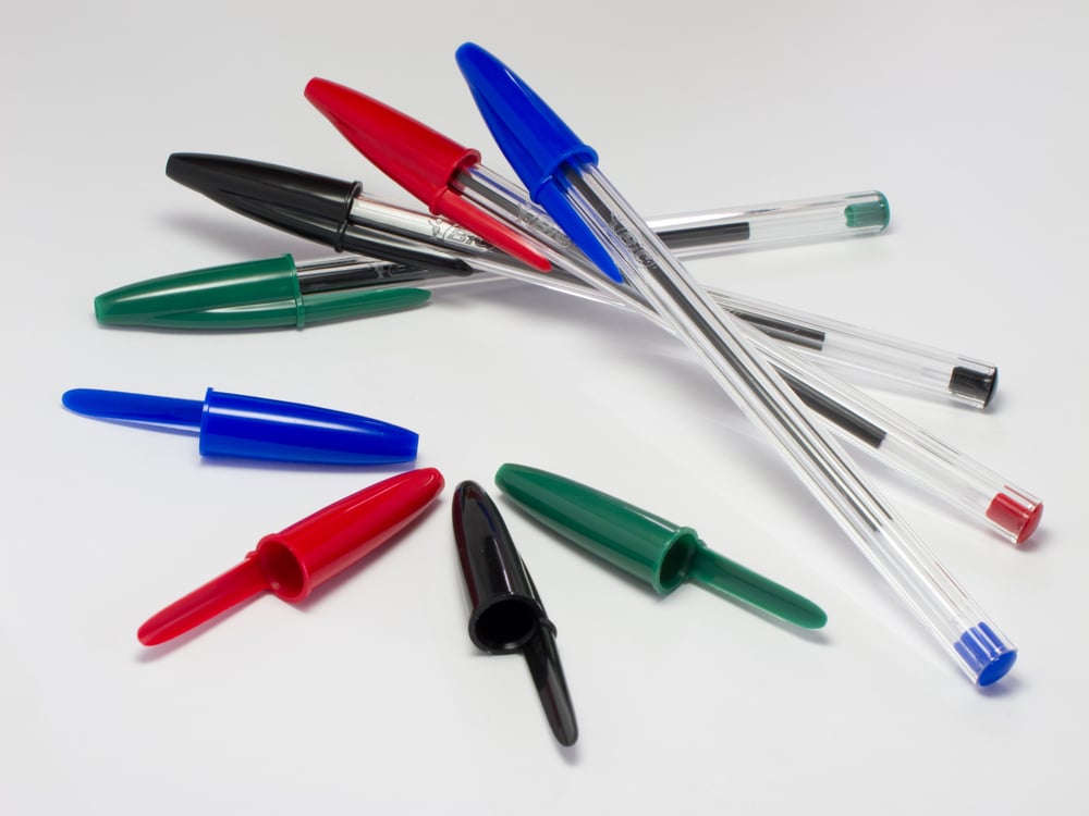 Several ballpoint pens with clear barrels and caps in red, blue, green, and black are scattered on a white surface. Some pens have their caps removed and placed nearby.
