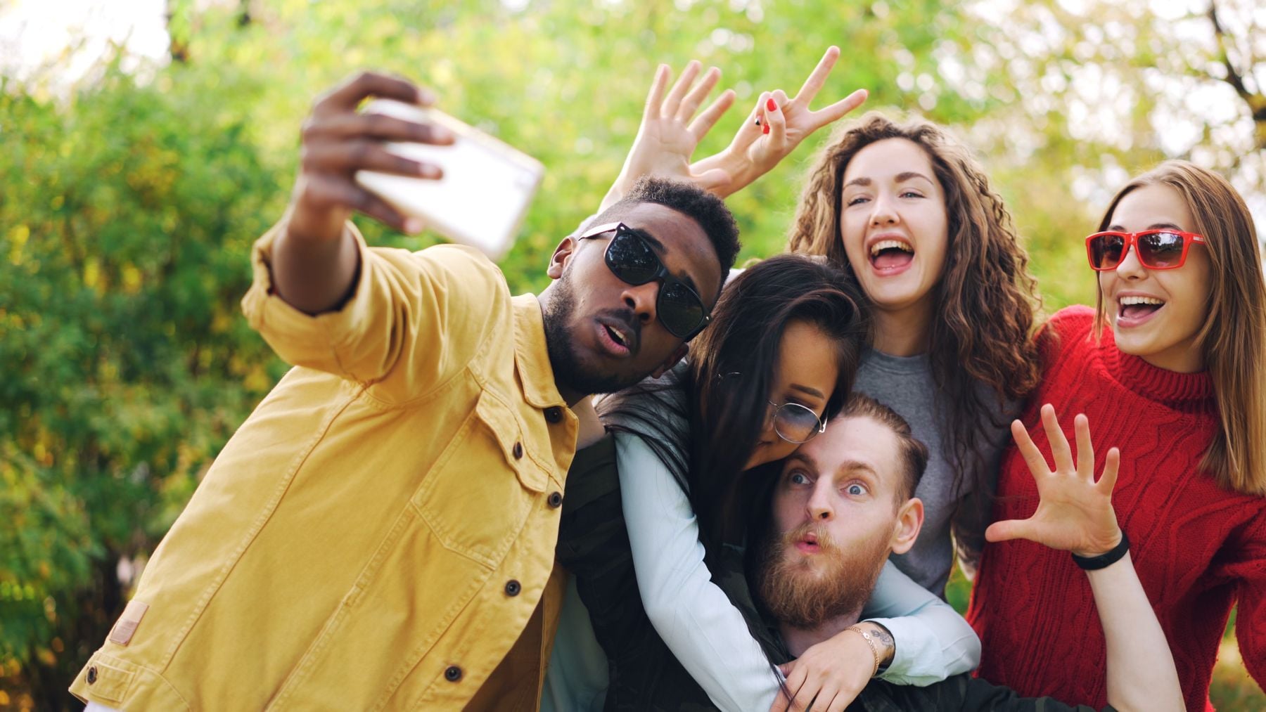 Five friends pose outdoors for a selfie, smiling and making playful gestures. The person in front wears sunglasses and holds the phone, while the others crowd together, looking cheerful. Trees are visible in the background.