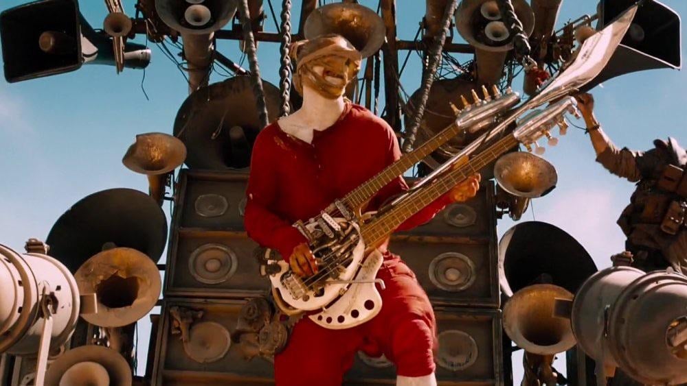 A person in red clothing and a face covering plays a double-neck guitar in front of a wall of speakers and amplifiers under a blue sky.
