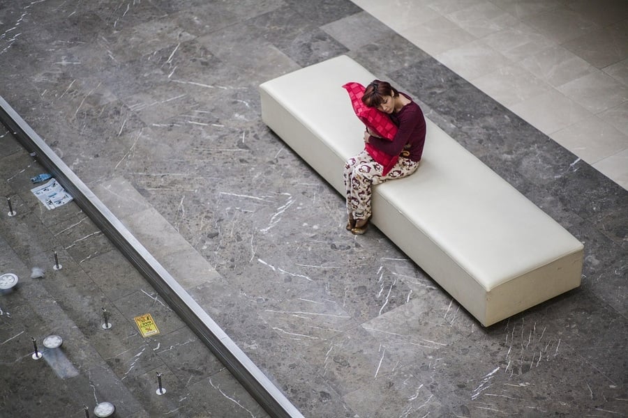 A person in patterned pants and a maroon top sits alone on a white bench, hugging a red pillow, on a gray marble floor in a spacious indoor area.