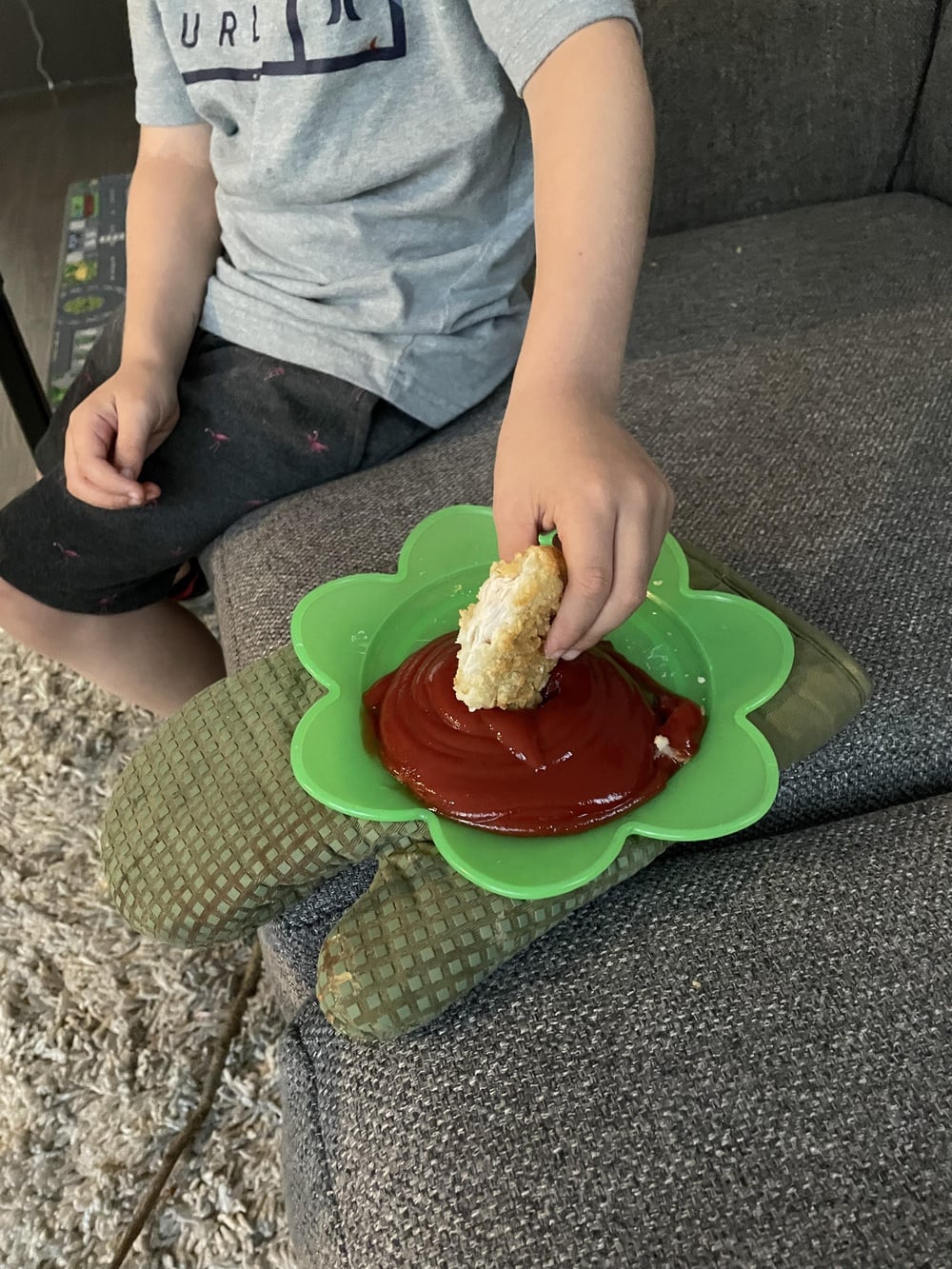 A child sits on a couch, dipping a breaded chicken tender into a large amount of ketchup on a green, flower-shaped plate placed on top of oven mitts.