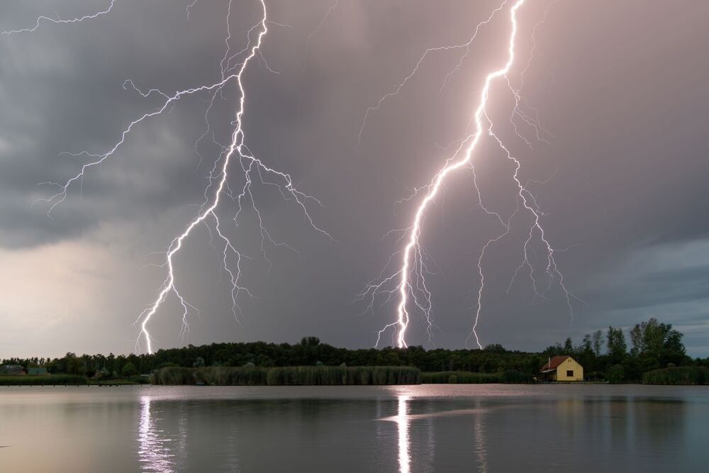 Three bright lightning bolts strike near a small yellow house by a lake, with their reflections visible in the water. The sky is gray and cloudy, creating a dramatic scene.
