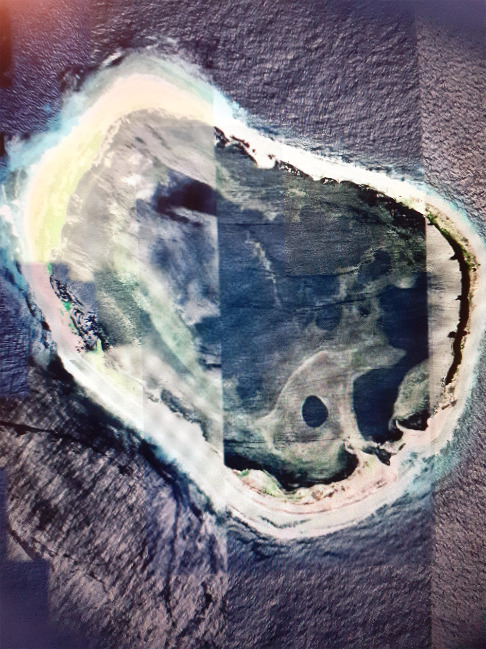 Aerial view of a small, oval-shaped island surrounded by blue ocean, with sandy beaches and green vegetation. The image appears to have been taken in overlapping sections.