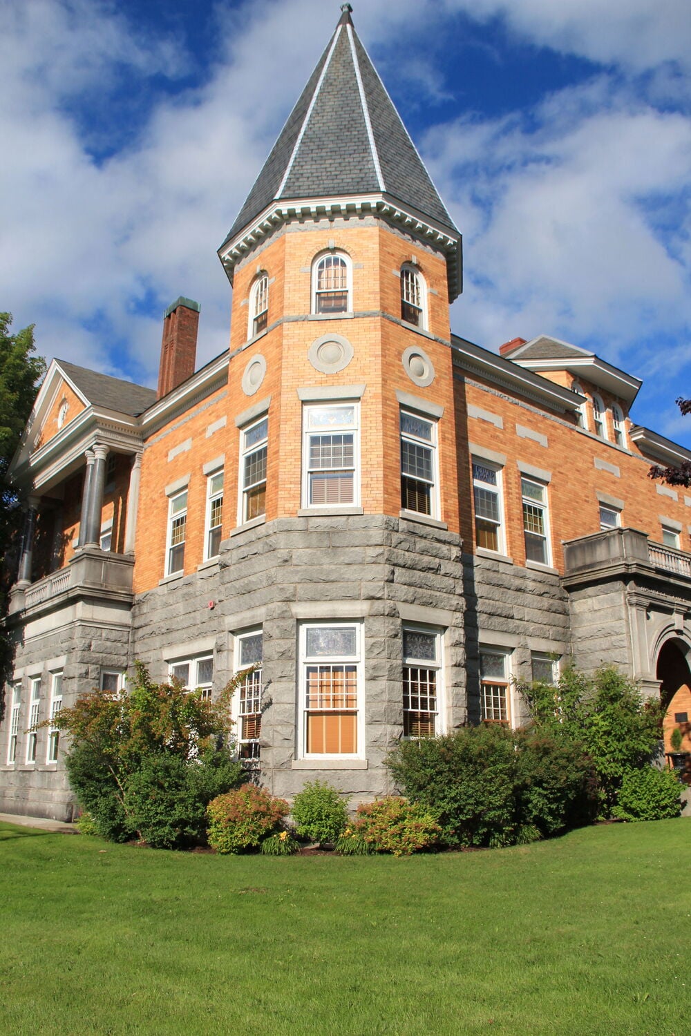 A historic brick and stone building with a tall, pointed tower, arched windows, and lush green lawn under a blue sky with scattered clouds.