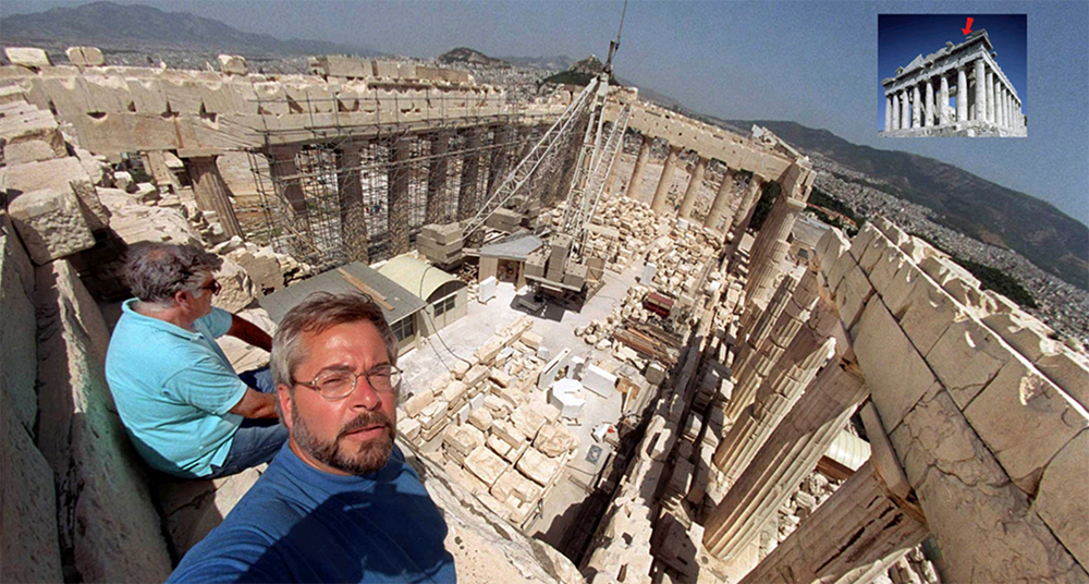 Two men sit on ancient stone blocks inside the partially restored Parthenon in Athens. Scaffolding and construction materials are visible, with hills in the background. An inset shows the Parthenon’s complete exterior.