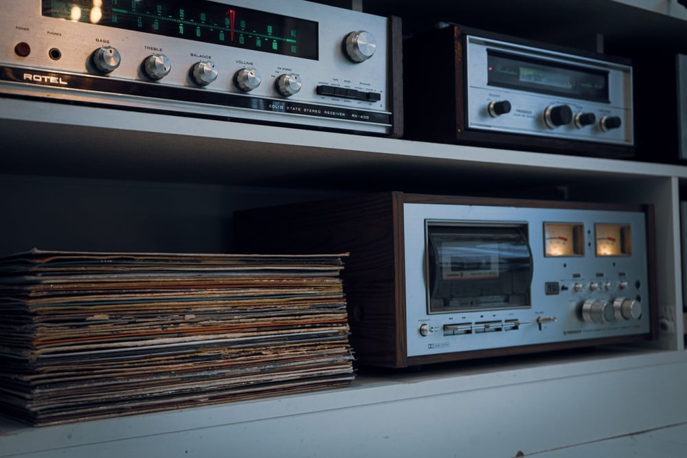 Vintage audio equipment, including a stereo receiver, cassette deck, and vinyl records, displayed on white shelves. The devices have metal finishes and knobs, evoking a retro aesthetic.