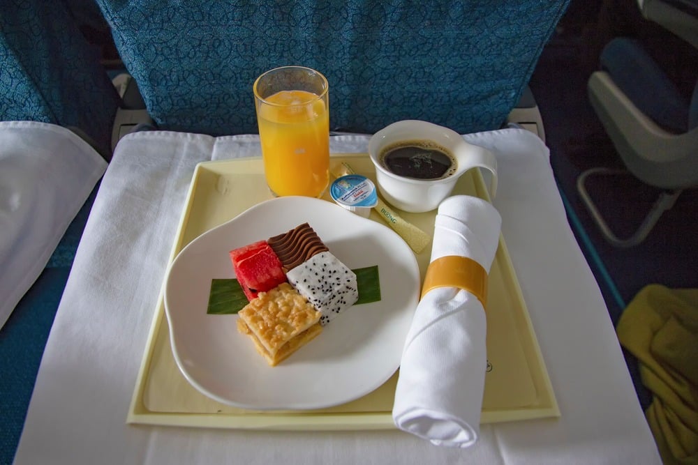 A tray on an airplane seat holds a plate of assorted desserts, a glass of orange juice, a cup of coffee, a butter packet, and a rolled napkin with a yellow band.