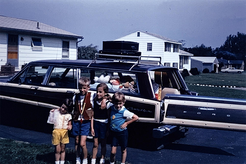 Four children stand smiling in front of a packed Ford station wagon with open rear hatch in a suburban driveway, houses and green lawn in the background.