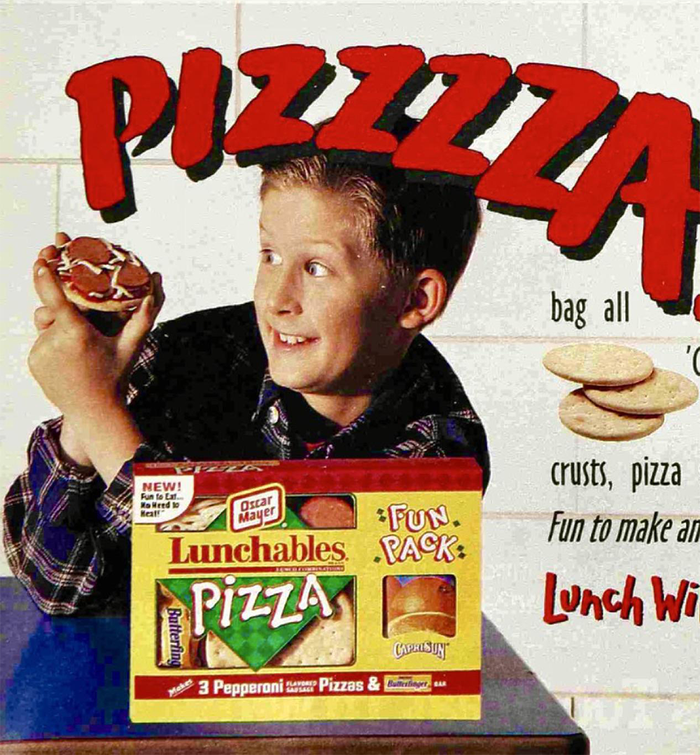 A smiling child holds a mini pizza with sauce, in front of a box of Lunchables Pizza. The word “PIZZZZA” is in large red letters in the background.