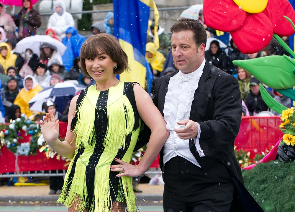 A woman in a yellow fringed dress and a man in a tuxedo dance together during a parade, surrounded by colorful decorations and a cheering, raincoat-clad crowd.