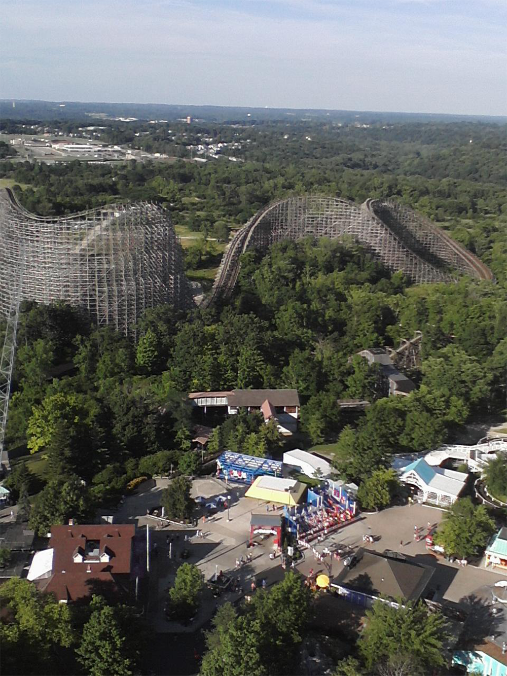Aerial view of a wooden roller coaster winding through trees, with a small fairground area and buildings visible below, surrounded by lush green forest and distant roads.