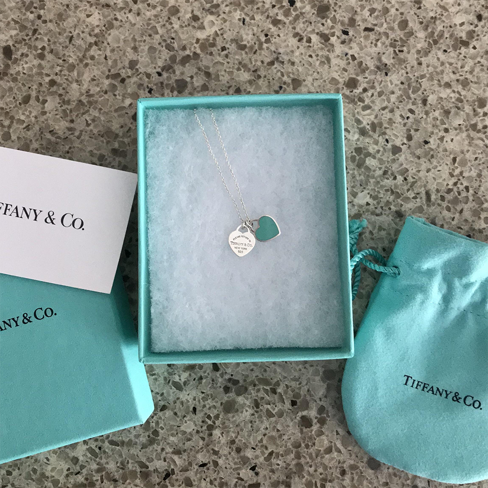 A Tiffany & Co. necklace with a silver heart and a teal heart charm in a blue box, next to a matching pouch and card, on a speckled surface.