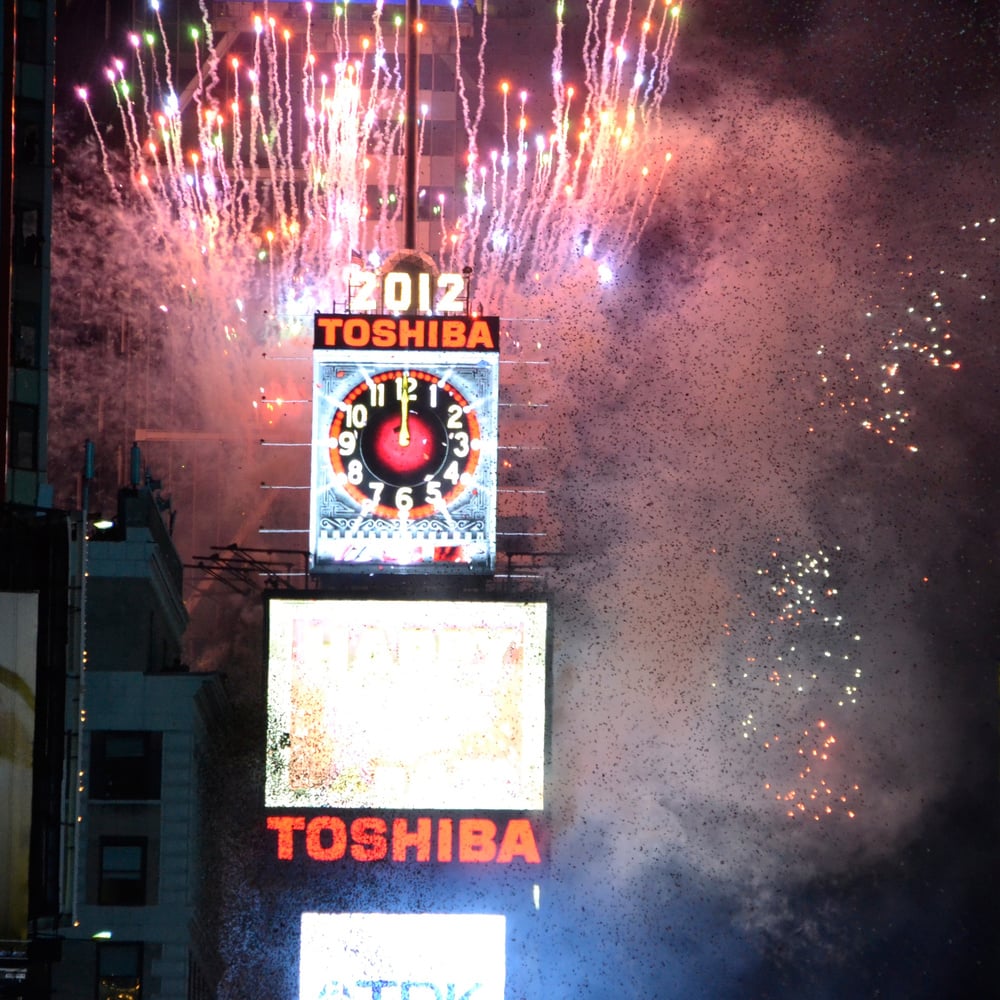 Fireworks explode over the Times Square New Year’s Eve Ball with “2012” and “Toshiba” signs lit up, marking the start of the new year amidst bright lights and confetti.