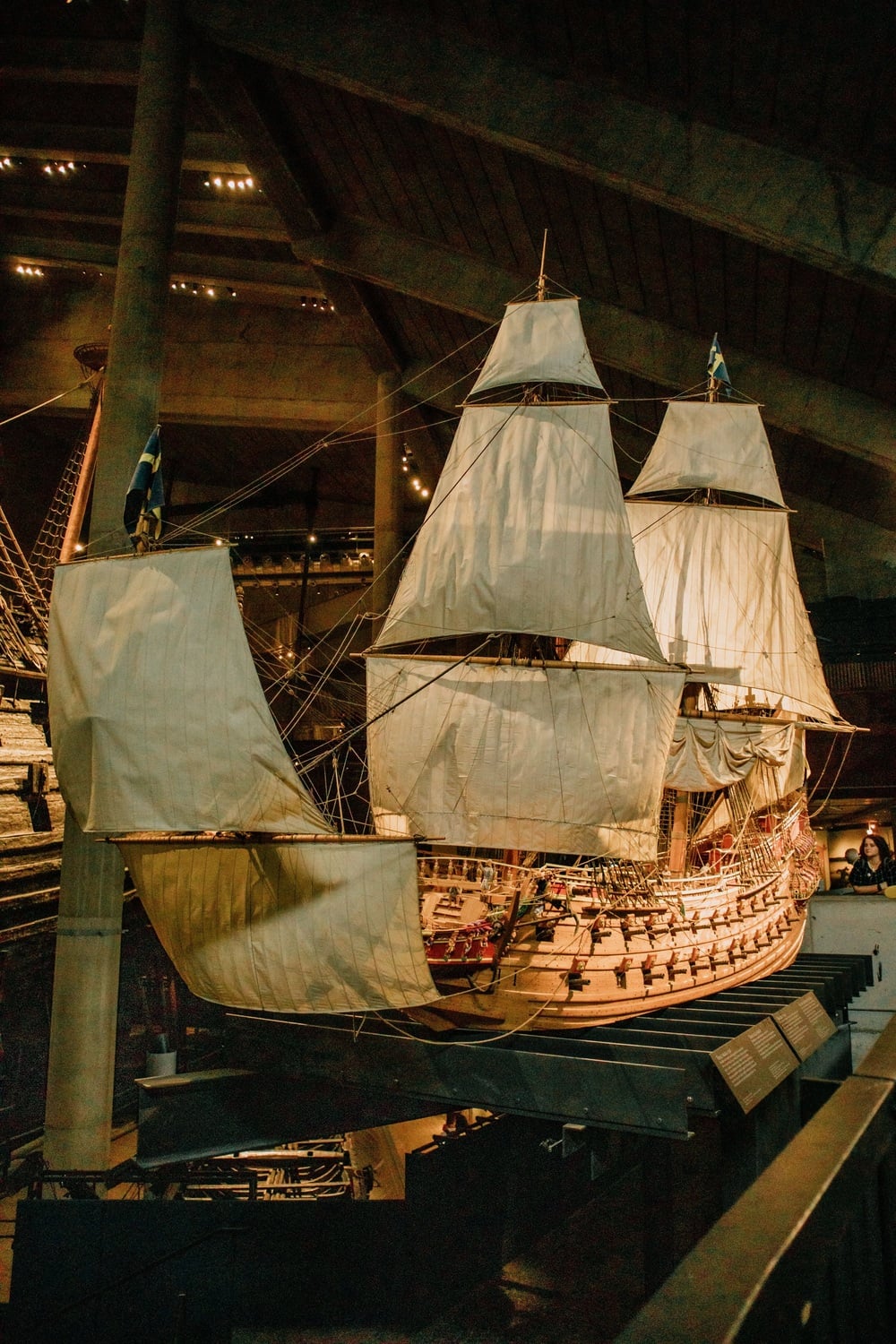 A large wooden sailing ship with white sails is displayed indoors at a museum, surrounded by railings and informational plaques.