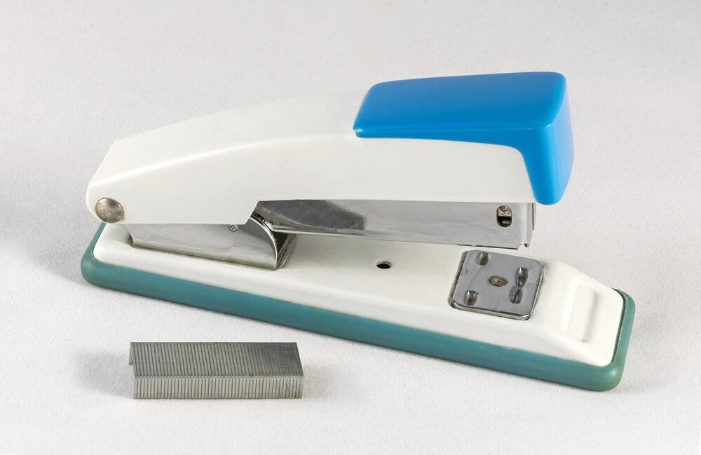 A white and teal stapler with a blue top is placed on a light surface, next to a small stack of unused metal staples.