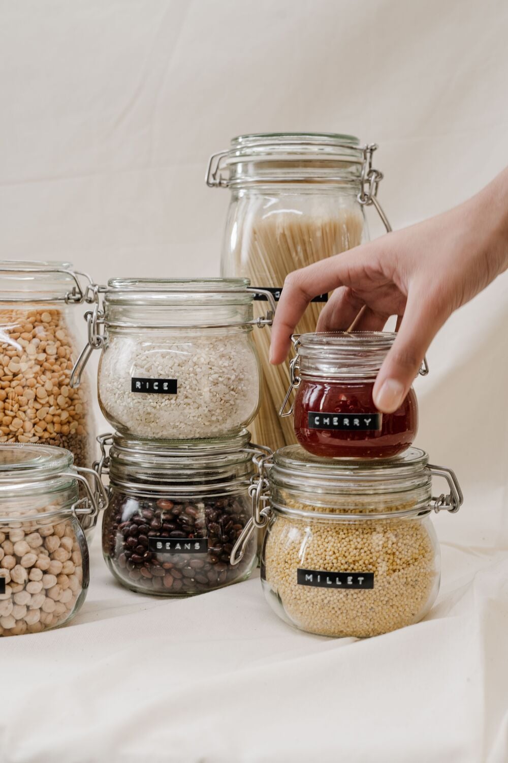A hand reaches for a labeled glass jar of cherries among jars of rice, beans, millet, chickpeas, lentils, and spaghetti, all organized on a light surface.