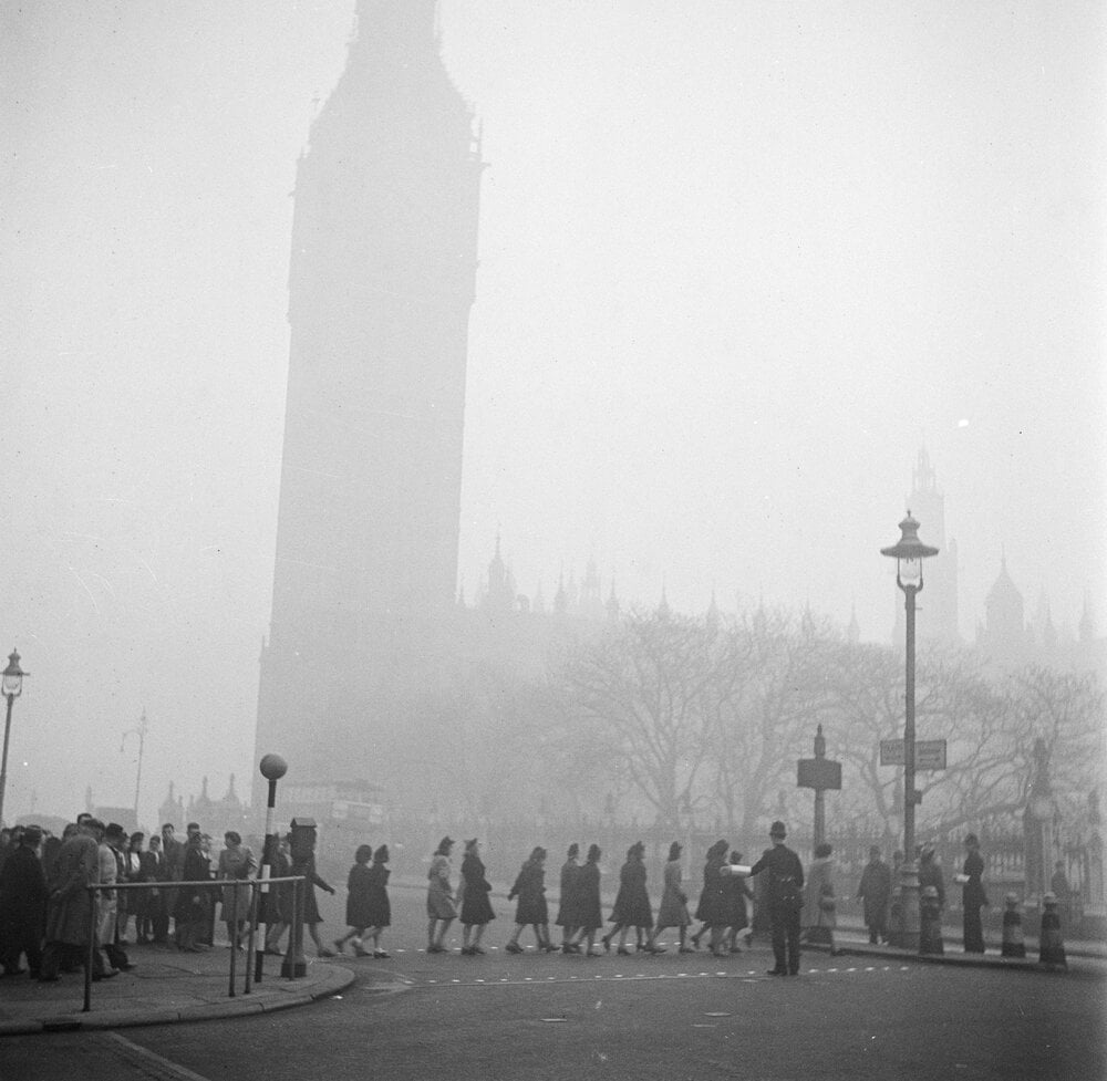 Pedestrians cross a foggy street near the Palace of Westminster and Big Ben in London; the scene is shrouded in thick smog, obscuring building details and creating a sense of historical London atmosphere.