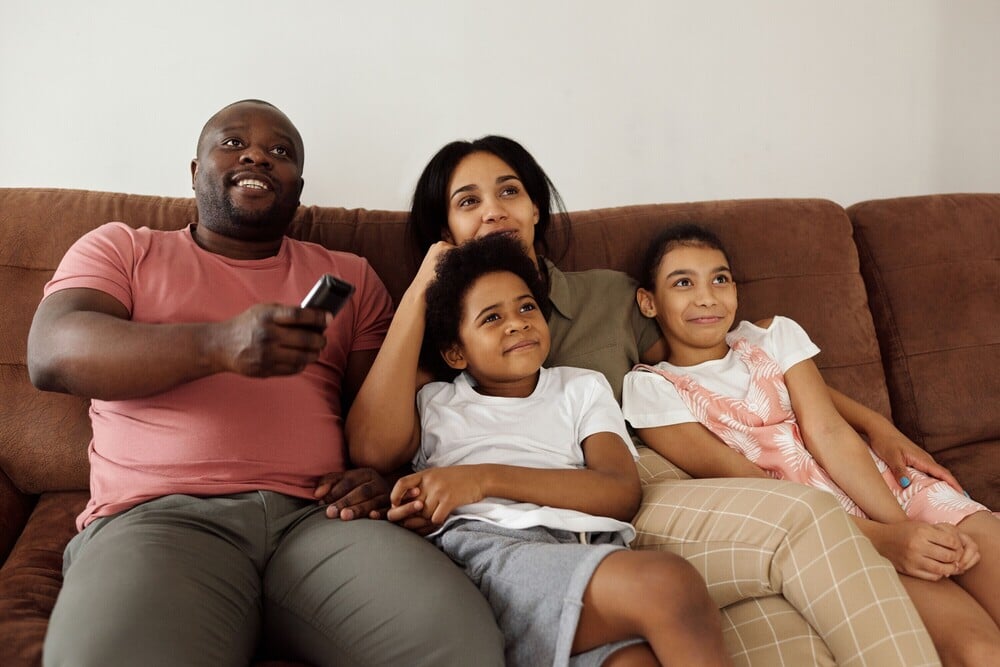 A family of four sits together on a brown couch, smiling and watching TV. The father holds a remote, while the mother and two children sit close, looking content and relaxed.