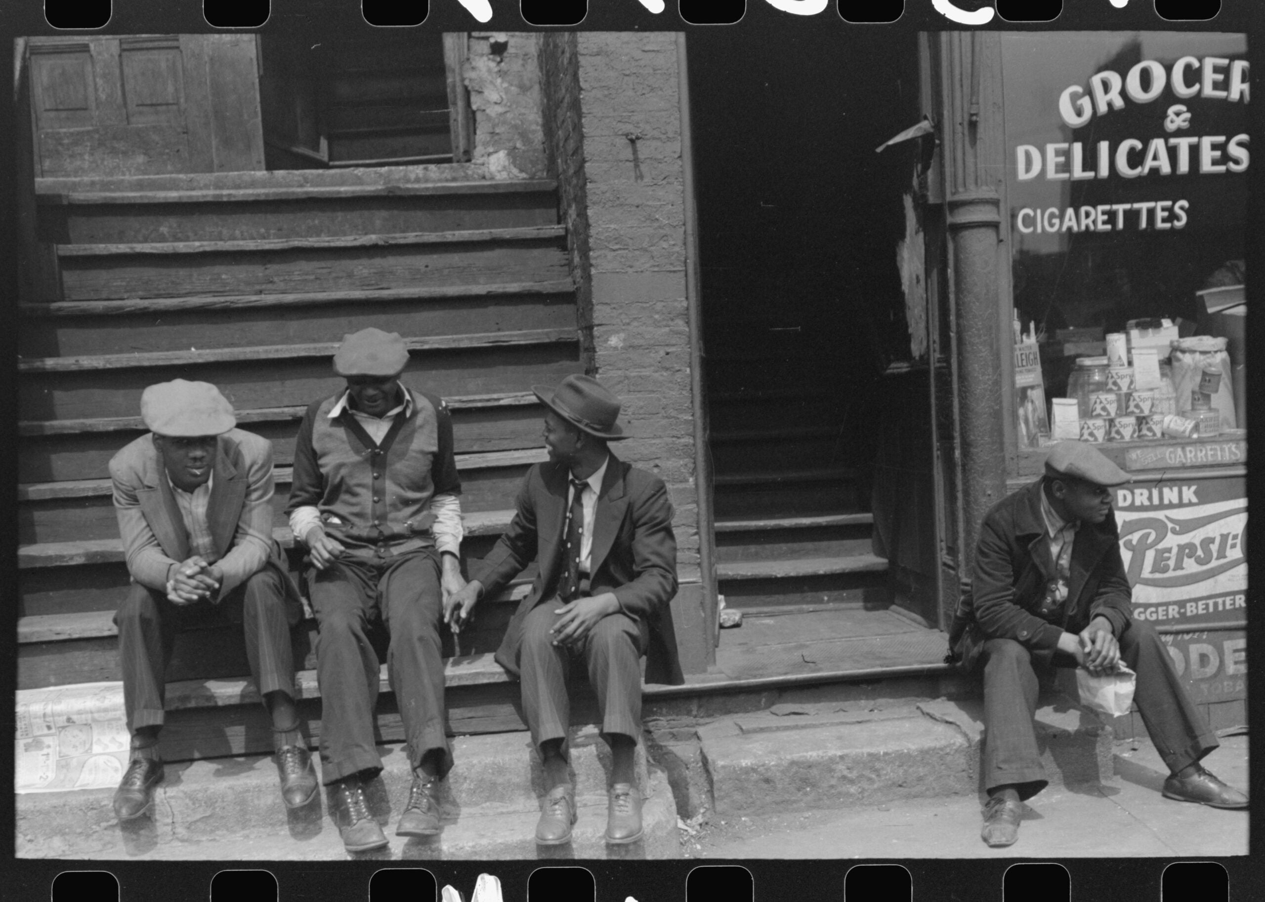Four men sit on steps outside a store with a “GROCER & DELICATES” sign. One man sits apart while the others talk. The storefront advertises Pepsi and cigarettes. The scene is in black and white.