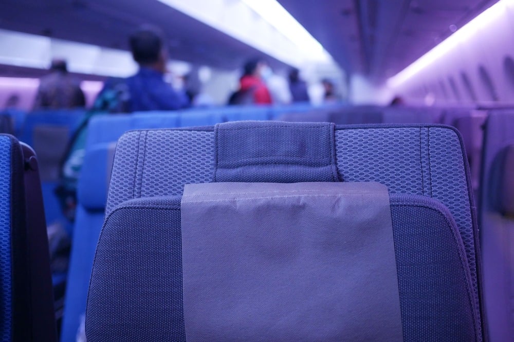 Close-up of a blue airplane seat with a headrest cover, showing rows of seats and blurry passengers in the background under purple cabin lighting.