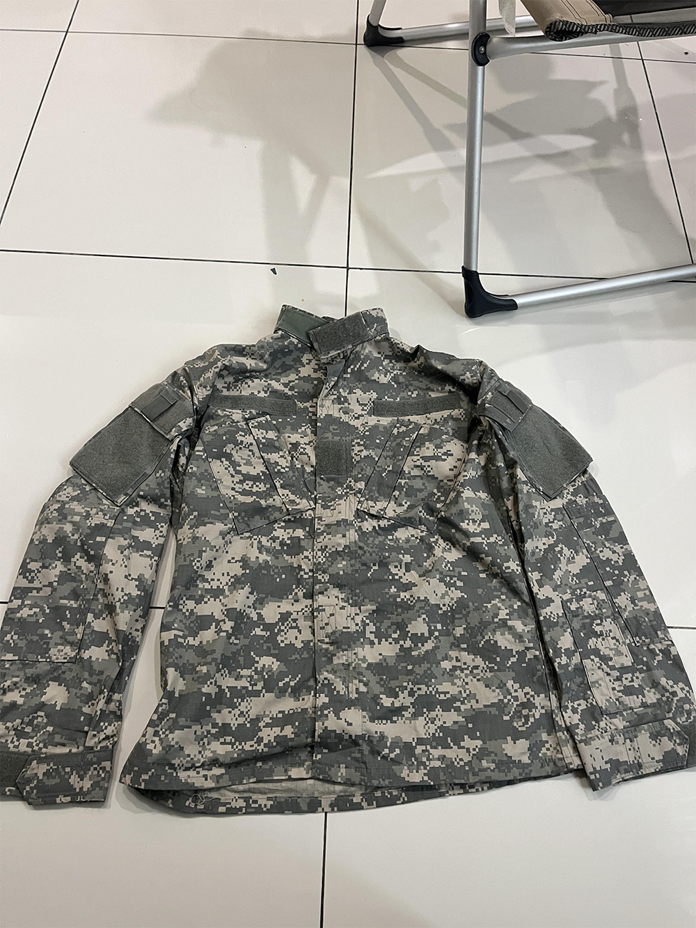 A digital camouflage military jacket lies flat on a white tiled floor next to a metal folding chair.