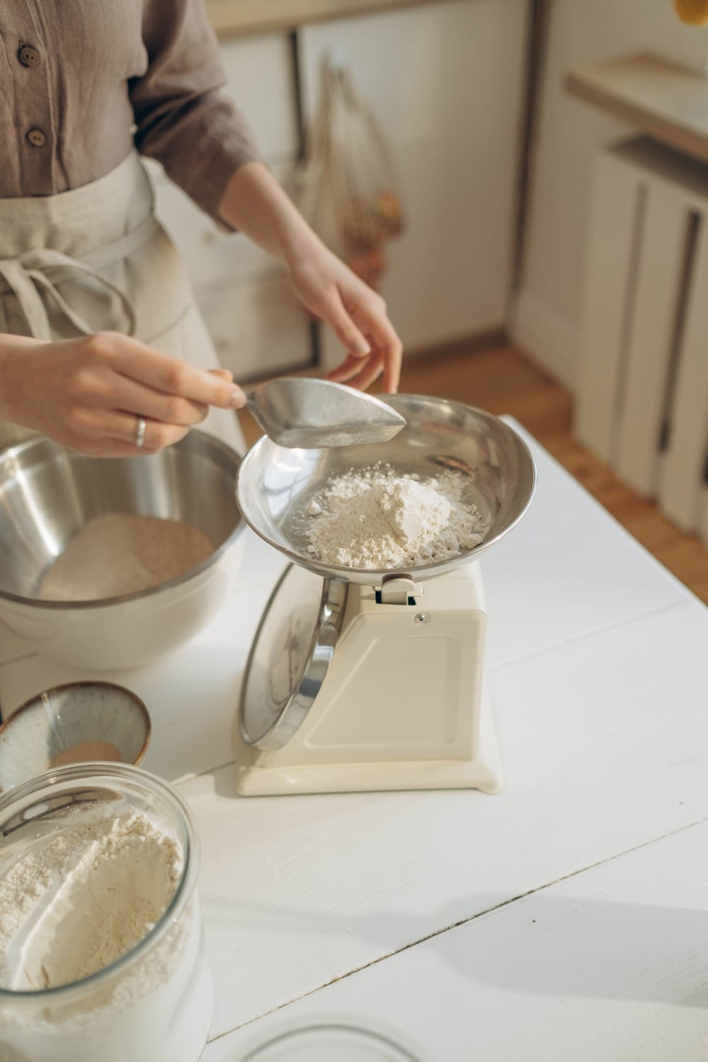 A person measures flour on a vintage kitchen scale, holding a metal scoop over the scale. Mixing bowls and a jar of flour are on the white countertop in a bright kitchen.