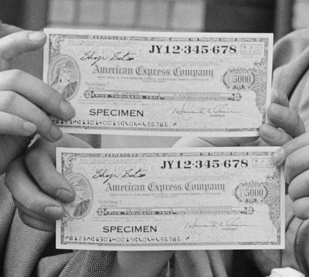 Two hands each hold an American Express Company $5,000 traveler’s cheque labeled “SPECIMEN,” with identical serial numbers, signatures, and detailed design elements visible.