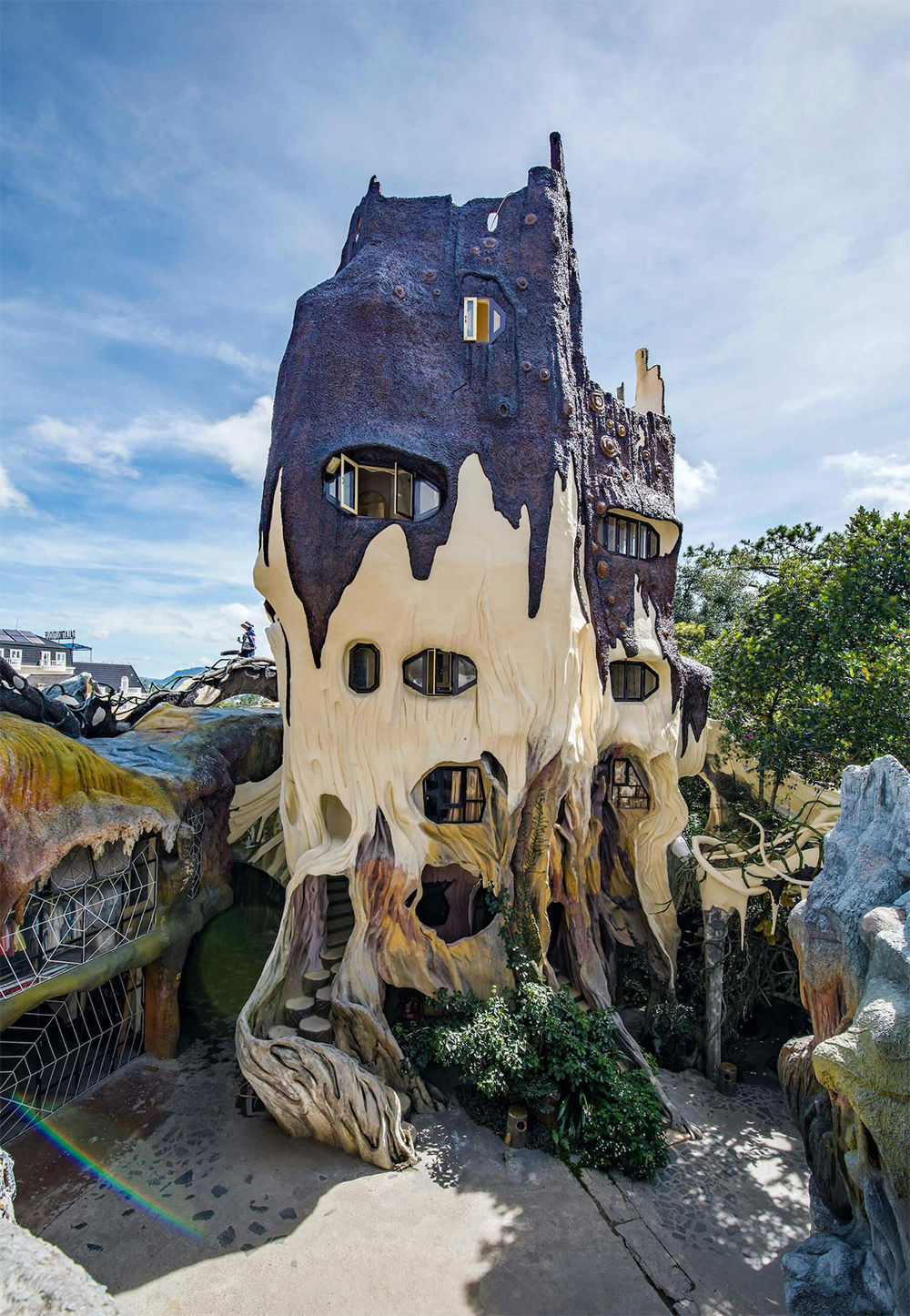 A whimsical, tree-like building with irregular windows and a melted, organic facade, surrounded by greenery under a blue sky.