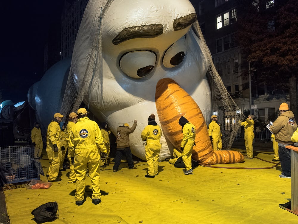 People in yellow suits prepare a giant snowman balloon, likely for a parade, at night on a city street. The snowman’s head is surrounded by workers adjusting or inflating it.