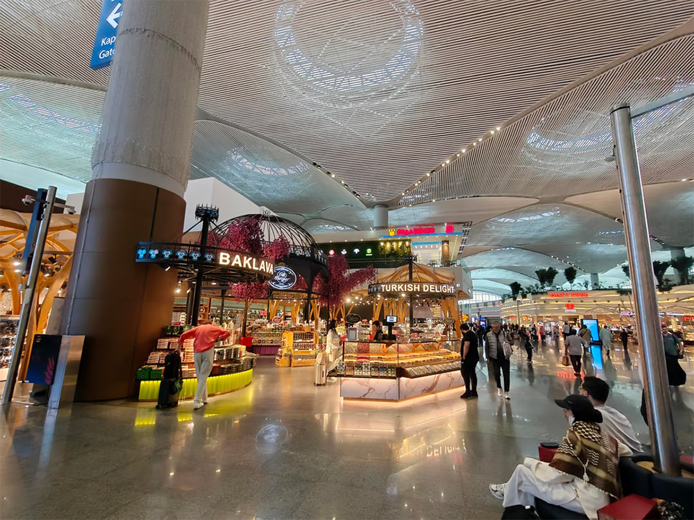 A bright, spacious airport terminal with shops selling baklava and Turkish delight. Travelers walk or sit, and the modern ceiling has intricate patterns. Signs and displays are visible throughout the scene.