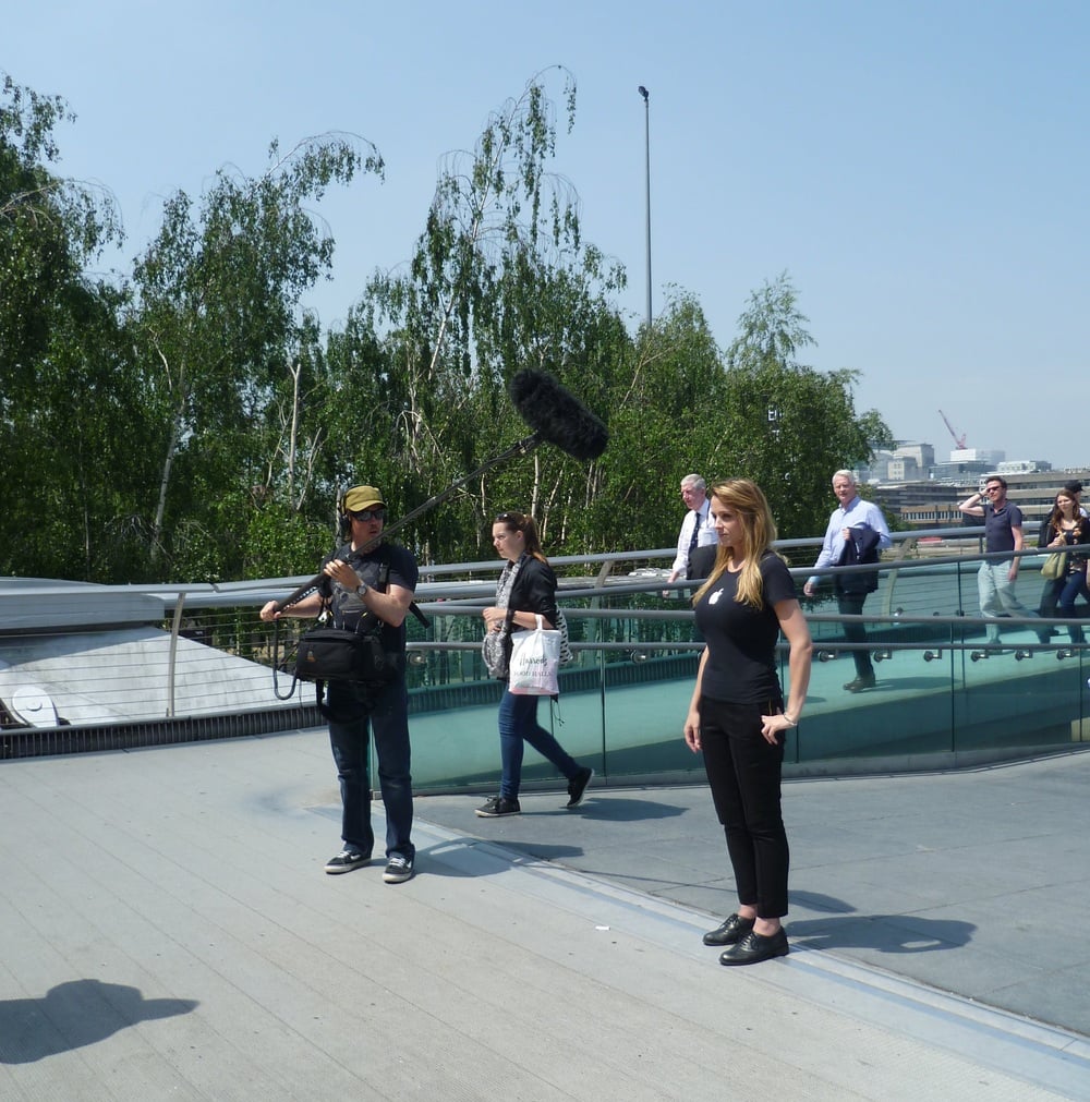 A film crew records a woman standing on a pedestrian bridge outdoors. One person holds a boom microphone while another operates a camera. Several people walk in the background amid trees and modern railings.