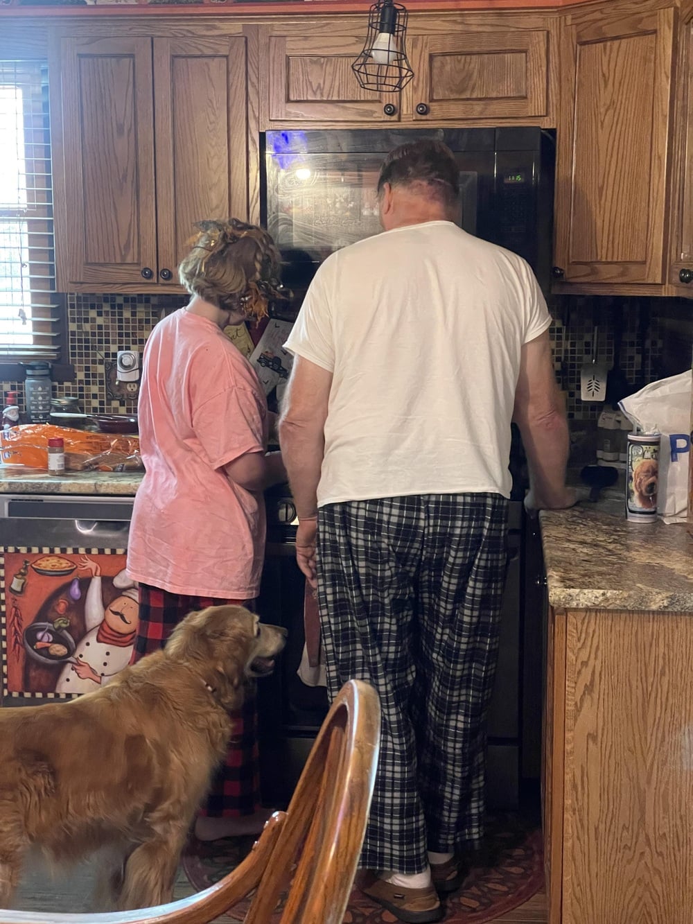 Two adults in pajamas stand in a kitchen near the fridge, with a golden retriever looking up at them. The kitchen has wooden cabinets and various food items on the counter.