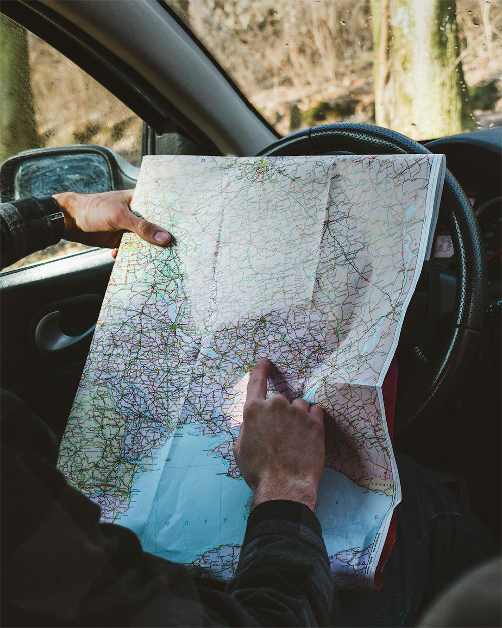 A person sits in a car, holding and pointing at a large paper map spread over the steering wheel, with sunlight shining through the window.