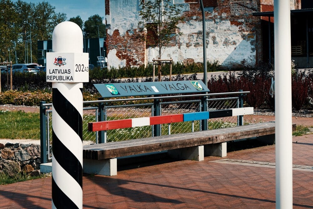 A striped border marker stands beside a bench labeled “Valka Valga,” representing the Latvia-Estonia border. A sign on the marker reads “Latvijas Republika.” Brick buildings and plants are visible in the background.