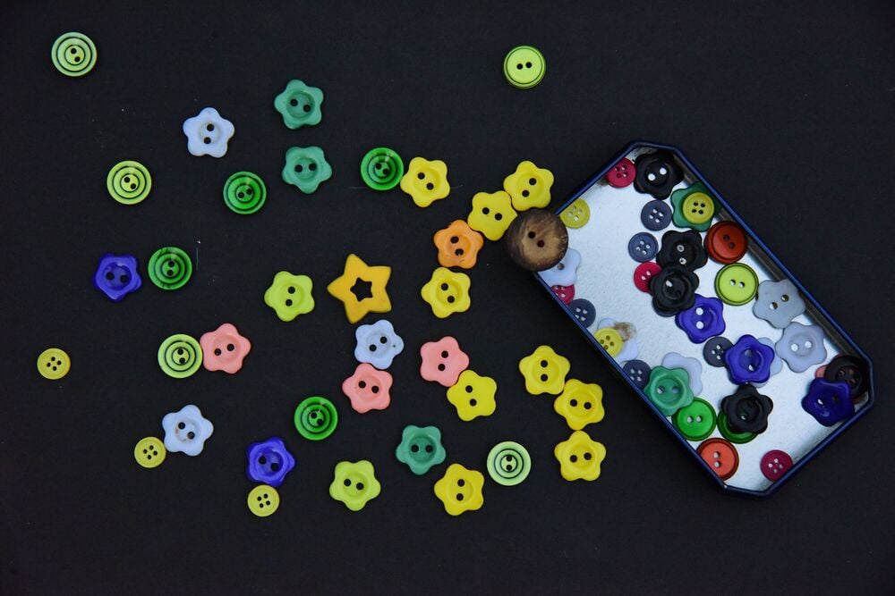 Colorful buttons, mostly flower-shaped, are scattered on a black surface, some spilling out of a rectangular tin container.