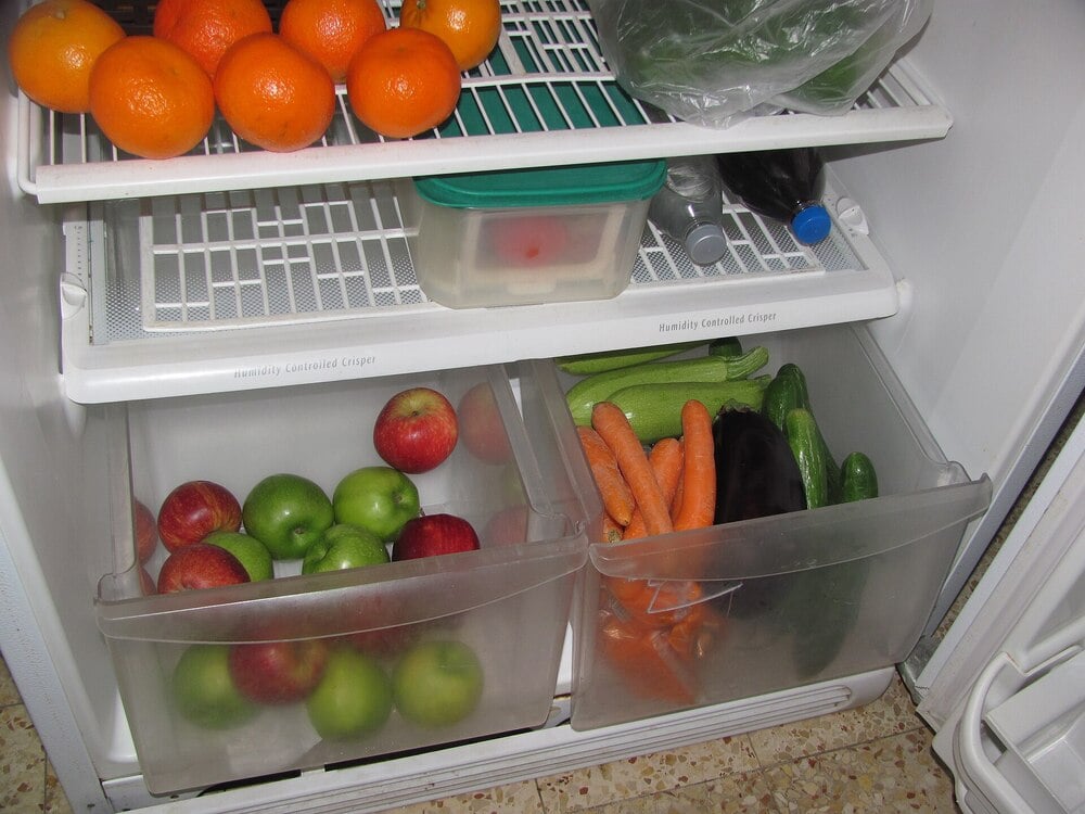 Open refrigerator with oranges, apples, carrots, cucumbers, an eggplant, a green plastic container, and a bottle on the shelves and in clear drawers.
