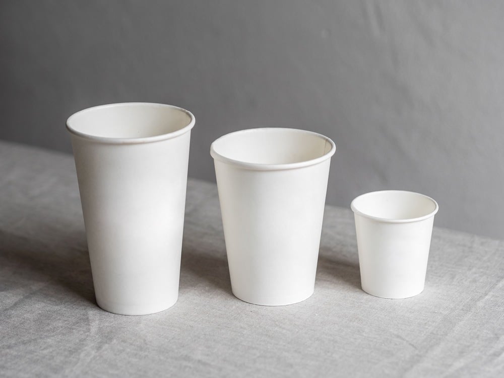 Three plain white paper cups of different sizes are arranged in a row on a light gray surface, against a plain gray background.